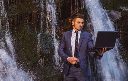 Attractive business man in suit holding laptop at waterfall background. Telework stressful work concept.の写真素材