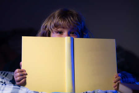 Close up face of child little boy reading a book in the dark home. Kids face with night light.の写真素材