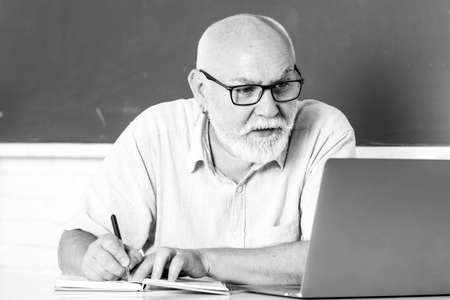 Elderly education and retired knowledge concept. Senior teacher teaching in the classroom on chalkboard. Teacher at university. Man senior teacher using laptop computer sitting at desk in classroom.の写真素材