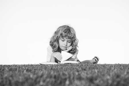 Smart child reading book, laying on grass in field on sky and grass field. Portrait of clever kids.