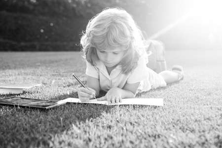 Kid boy reading a book lying on grass writing notes in copybook. Cute little child in casual clothes reading a book in park.の写真素材