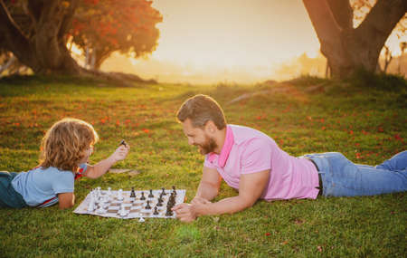Son laying on grass and playing chess with father on sunset in sunny park.の写真素材