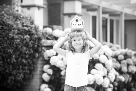 Real estate, housing and family concept. Funny kid holding toy wooden house near new home.の写真素材
