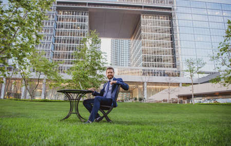 Businessman typing on laptop computer sitting at table on office terrace. Man working remotely in modern park.の写真素材