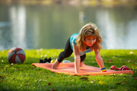 Child is pushing up on the green meadow, working out wearing sportswear, t-shirt outdoor. Kids doing...