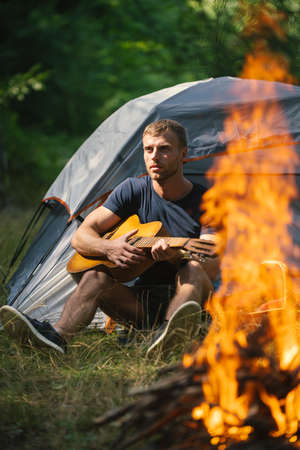 Young man sitting near touristic tent and playing guitar. Romantic time in a mountain camp. Tourism...