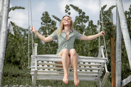 Outdoor portrait of a smiling happy girl. Fashion woman on wooden bench. Beauty blonde girl relaxing outdoors on porch swing in the nature. Summer mood. Happy cheerful girl laughing at park.の写真素材