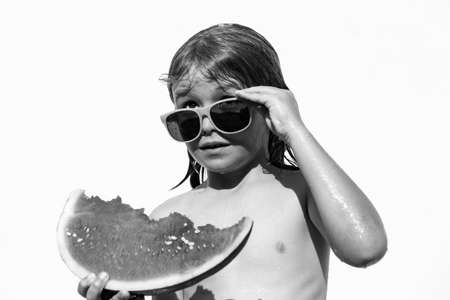 Kid eats watermelon in the pool, isolated on white. Cute boy eating slice of red watermelon on the beach, happy caucasian boy watermelon against the blue water.の写真素材
