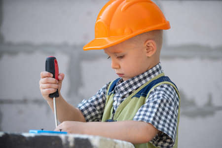 Kid boy twists bolt with screwdriver. Child repairman with repair tool. Child in helmet and...