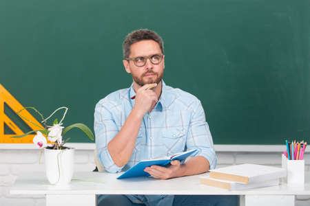 Man teacher thinking. Portrait of smart young tutor in glasses with book think on the blackboard in class at school or collage. Male student study in university.の写真素材