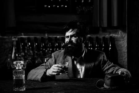 Handsome pensive man is holding a glass of whiskey and looking away while sitting in bar indoors.