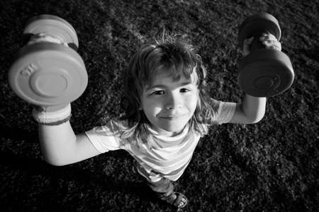 Funny child sport. Kid with dumbbells in park. Strong little boy. Fun child face. Children development and healthy strong exercise. Wide angle.の写真素材