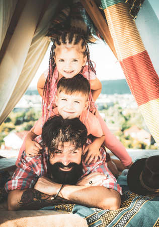 Young handsome father spend his free time with kids. Children put their heads in one vertical row with their fathers head. Little pyramid house at the roof for having fun with family.の写真素材