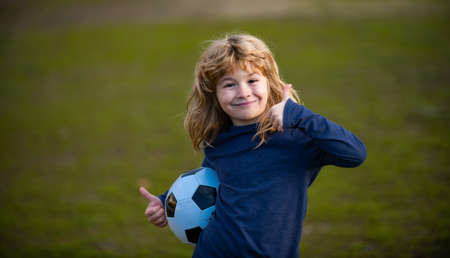 Boy child playing football on football field. Kid playing soccer show thumbs up success sign. Boy holding soccer ball, close up sporty kids portrait.の写真素材