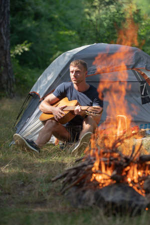 Young man sitting near touristic tent and playing guitar. Romantic time in a mountain camp. Tourism and hike, travel lifestyle. Sexy man playing guitar in forest with bonfire.の写真素材