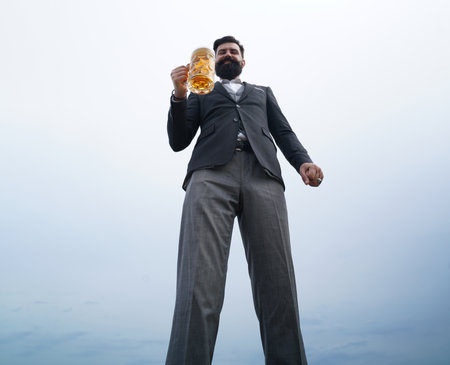 Funny bearded drunk hipster man holds craft beer on sky nature background. Full length, wide angle,...