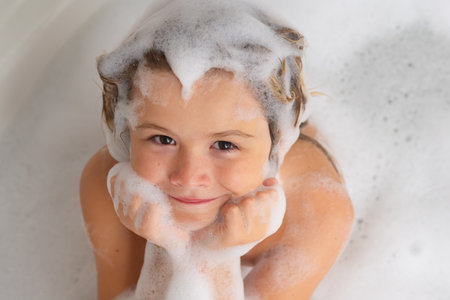 Kids shampoo. Child with shampoo foam and bubbles on hair taking bath. Funny face of kid in foam,...