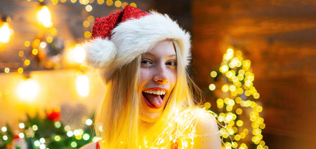 Funny woman in Santa hat on christmas background. Close up portrait of sexy girl flirting, show...