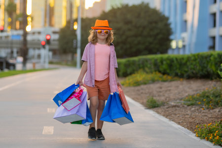 Cute little boy in summer fashion clothes goes shopping. Happy child with shopping packages in hands. Shopper child with shopping bag outdoor. Cute kid walking on street carrying shopping bags.の写真素材