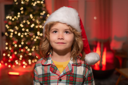 Child in santa hat on front of night Christmas tree home background on Christmas Eve.