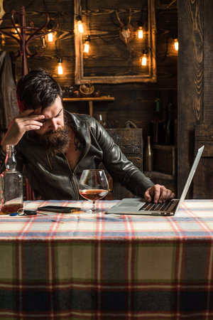 Hipster with beard and mustache drinks alcohol after working day. Under pressure. Businessman drinking cause of stress, holding glass of whiskey used laptop home interior.の写真素材