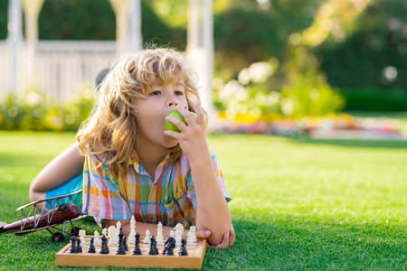 Chess school outdoor. Child think or plan about chess game, laying on grass in summer park, eating apple. Intelligent, smart and clever school kids.の写真素材