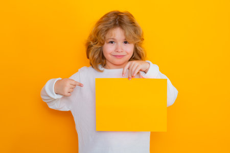 Cute child showing index finger on sheet of paper, isolated on yellow background. Portrait of a kid...