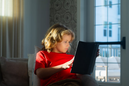 Cute kid in casual clothes reading a book while lying on a sofa in the room. Smart, clever child....