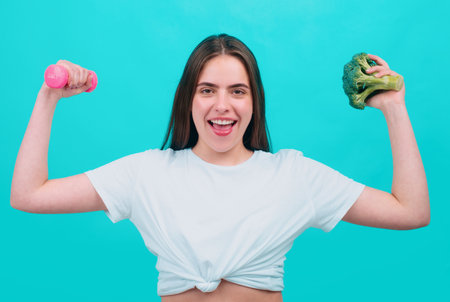 Portrait of young beautiful woman with broccoli and dumbbell, over blue background. Healthy food,...