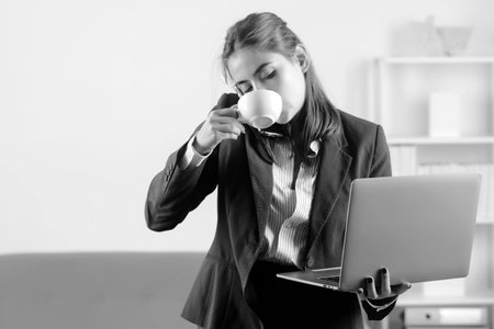 Young businesslady at workplace in the office. Secretary woman in formal wear drinking coffee, talking on phone, using laptop computer.の写真素材