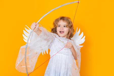 Angel child shoots a love arrow from a bow on Valentines Day. Valentines day. Kid with angel wings. Studio portrait of angel child on studio color isolated background with copy space.の写真素材