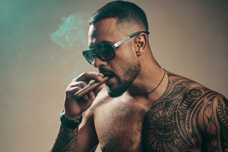 Attributes of men power, strength and luxury life. Strong confident tattooed man in luxury sunglasses smoking an elite cigar isolated at gray background. Bearded careless man smoke.の写真素材