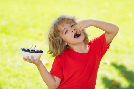 The child eats cherries. Excited face. Cherry for kids. Teenager child hold plate cherries. Amazed expression, cheerful and glad. Summer background with green grass.の写真素材