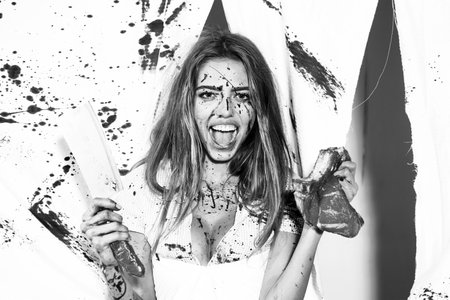 Dirty in blood, a woman holds in her hand a bloody cleaver on a white background in the studio. Young butcher girl holding a piece of raw meat and a cleaver isolated on bloody white background.の写真素材