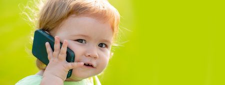 Baby on spring grass field, banner. Cute baby face closeup on green grass in summertime. Funny little kid portrait with mobile phone, smartphone on nature. Happy Childhood.の写真素材
