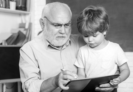 Portrait of confident old male teacher. Happy cute clever boy and old tutor with book. Educational process. Friendly child boy with old mature teacher in classroom near blackboard desk.の写真素材