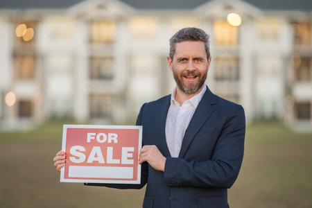 Real estate agent hold house sale sign. Real estate sale or property investment concept. Buying new home. Handsome real estate agent in suit showing the house for sale.の写真素材