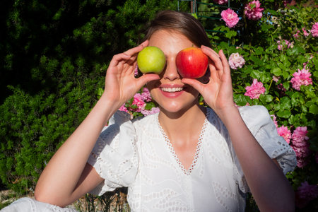 Happy young girl with two apples near her eyes. Woman hold tasty apple. Healthy teeth, girl holding apples near face in summer park. Dentistry, dentist and dental care concept.の写真素材