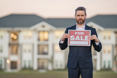 Realtor agent is a realtor with sign for sale in hand against the background on new apartment home background. Realtor in suit, outdoor portrait. Realtor offering new home. Property concept.の写真素材