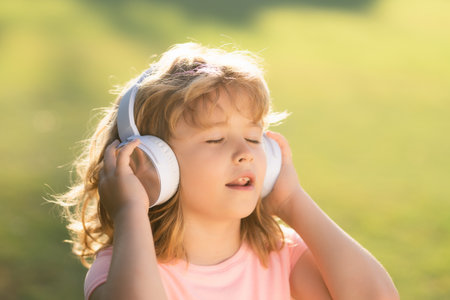 Singing children. Stylish teen boy listening music in headphones and singing against green grass summer background. Portrait of kid in headphones listening music with closed eyes.の写真素材
