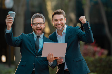 Two excited business men team using laptop outdoor. Amazed businessmen looking laptop with their business success in the city background. Two handsome young businessmen in classic suits using laptop.の写真素材