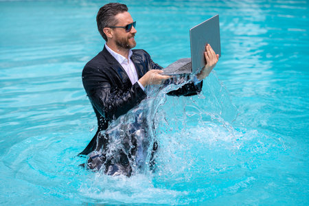 Businessman in suit with laptop in swimming pool. Crazy business man on summer vacation. Excited businessman in wet suit in swim pool. Funny business man, crazy comic business concept. Remote working.の写真素材