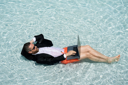 Funny businessman in suit working with computer on the sea beach. Summer holiday, outdoor and business travel concept. Crazy office worker. Man working on laptop computer sitting in cloth at poolside.の写真素材