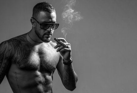 Expensive tobacco. Cigar smoking enjoy life moment. Bearded muscular macho beautiful torso fashion sunglasses smoking cigar. Elite tobacco. Pure enjoyment. Consuming tobacco. Tattooed man with cigarの写真素材