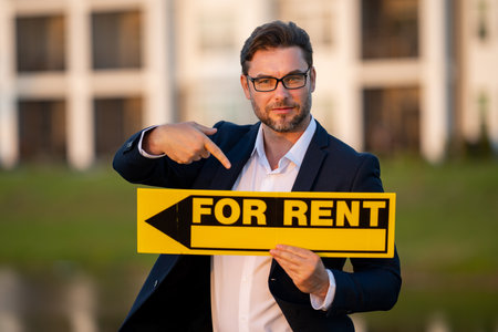 Successful real estate agent in a suit holding for rent sign near new apartment. Real estate agent with home loan contract, renting home. Realtor or real estate agent shows board for rent.の写真素材