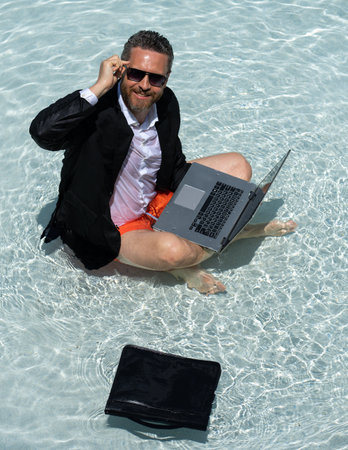 Business man in suit working on laptop in swimming pool. Travel tourism and business concept. Crazy male office employee using laptop in pool on summer day. Office conditioner concept.の写真素材
