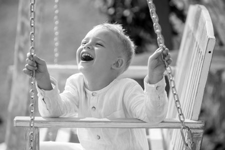 Happy child boy laughing and swinging on a swing at the park in summer. Kids happiness and smile. Child playing on outdoor playground. Summer kids portrait. Kid emotions concept.の写真素材