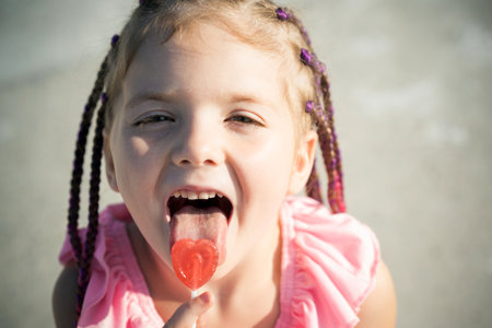 Little girl lick lollipop. Lifestyle portrait of cute kid outdoors. Summer kids outdoor portrait. Close-up face child playing outdoors in summer park. Kid having fun outdoor on sunny summer day.の写真素材