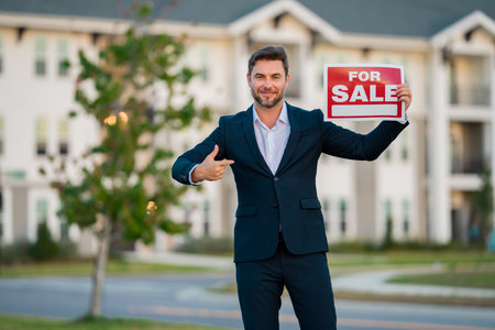 Successful real estate agent in a suit holding for sale sign near new apartment. Real estate agent with home loan contract, selling home. Realtor or real estate agent shows board for sale.の写真素材