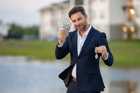 Successful real estate agent in a suit holding money dollars and keys from new apartment. Real estate agent with home loan contract, selling home. Man feeling happy about buying a new house.の写真素材
